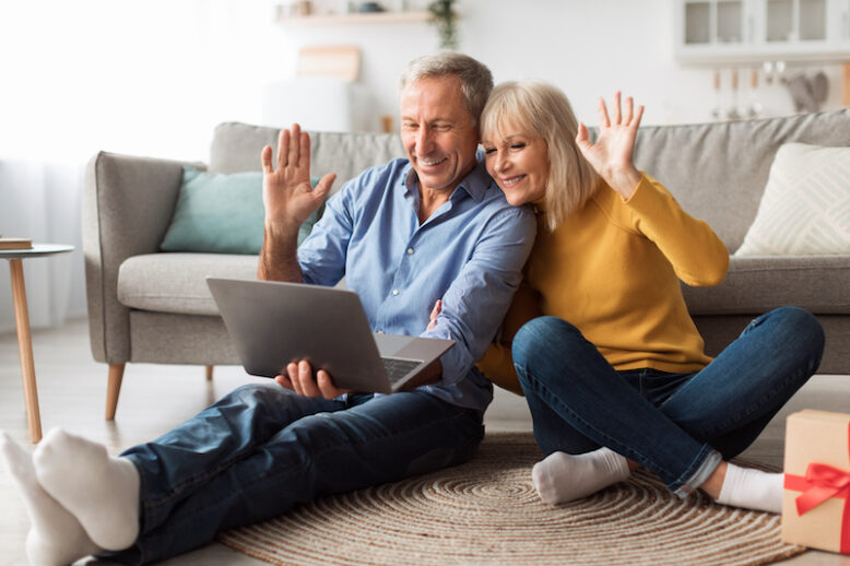 Senior Couple Video Calling Waving Hello To Laptop Webcam Communicating Online With Distance Family Members Sitting On Floor In Living Room At Home. Remote Communication Concept