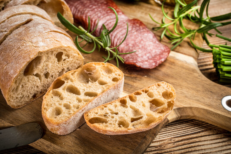 Italian ciabatta bread cut in slices on wooden chopping board with salami.