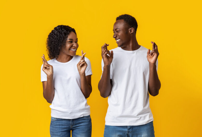 Making cherished wish. Young cheerful african-american couple with crossed fingers looking at each other and smiling, yellow studio background. Emotional black man and woman praying for good