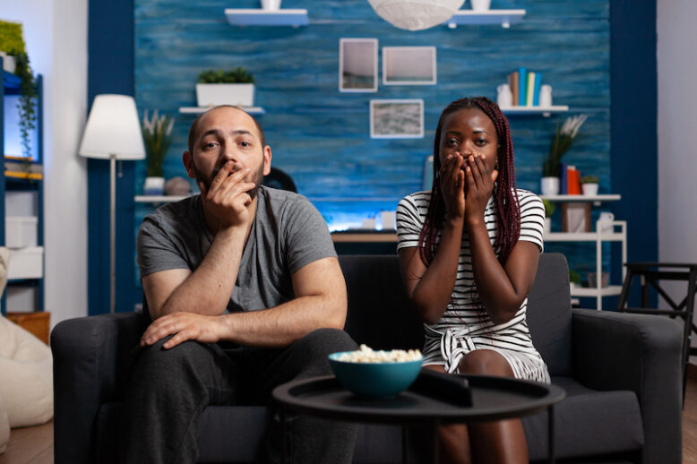 Shocked interracial couple watching tragic news on TV while sitting together at home on couch. Worried young people reacting to thriller movie on television program while sitting in living room