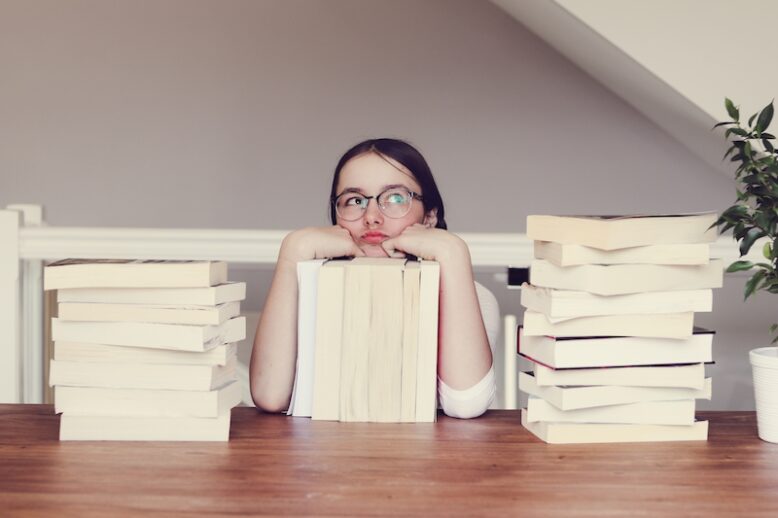 Cute funny tween girl in glasses sitting a table with pile of books looking sceptically and daydreaming of holidays. Student or pupil exhausted from reading