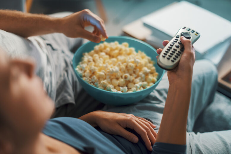 Couple sitting on the couch and watching TV, the woman is holding the remote control and the man is eating popcorn