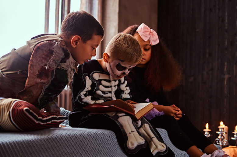 Three kids in scary costumes reading horror stories while sitting on bed in an old house.