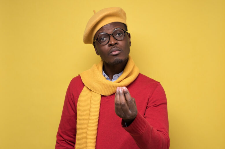 Young man in scarf and hat showing italian gesture that means what do you want over yellow background.