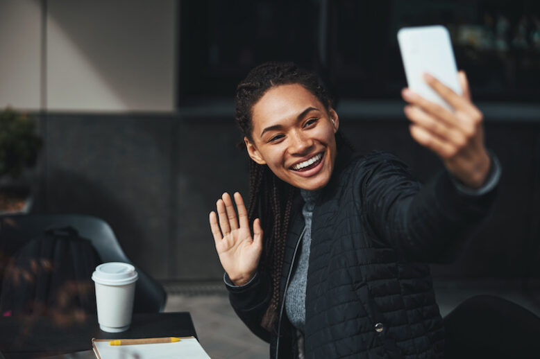 Lady holding a modern smartphone and waving to the camera while having an online meeting