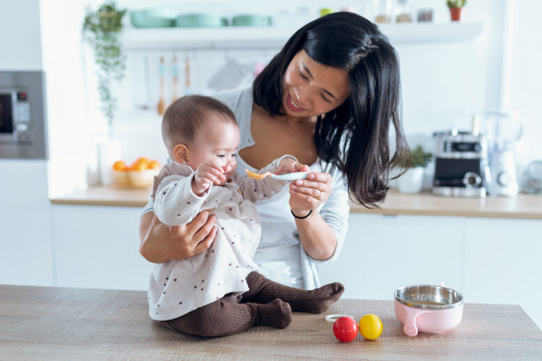Happy young mother feeding her cute baby girl with fruit porridge in the kitchen at home.