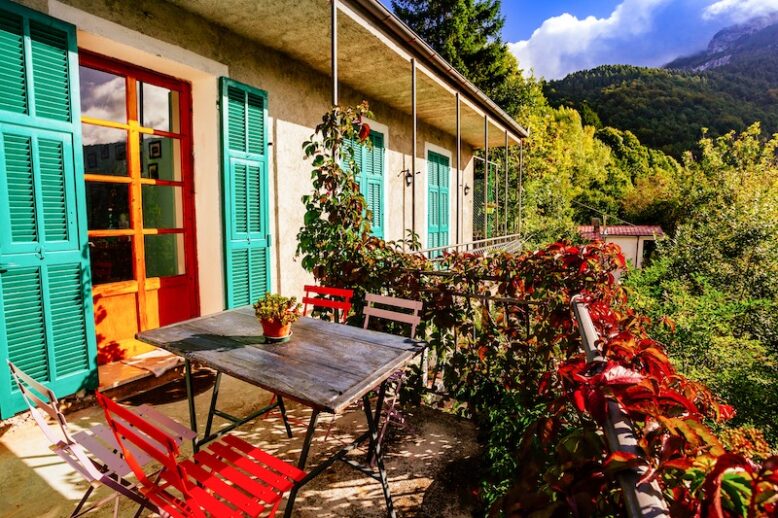 Traditional Italian house in the Dolomites in autumn colors. Patio with a vintage table and red chairs, turquoise window shutters.