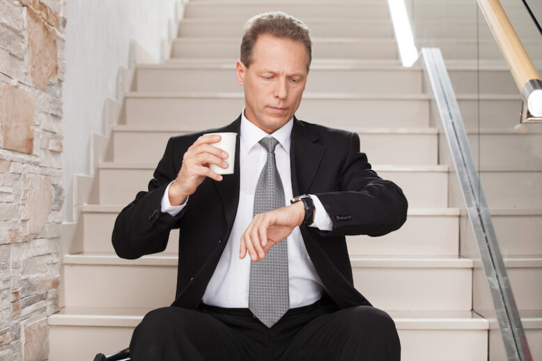 Worried mature man in formalwear holding a coffee cup and checking a time while sitting on staircase