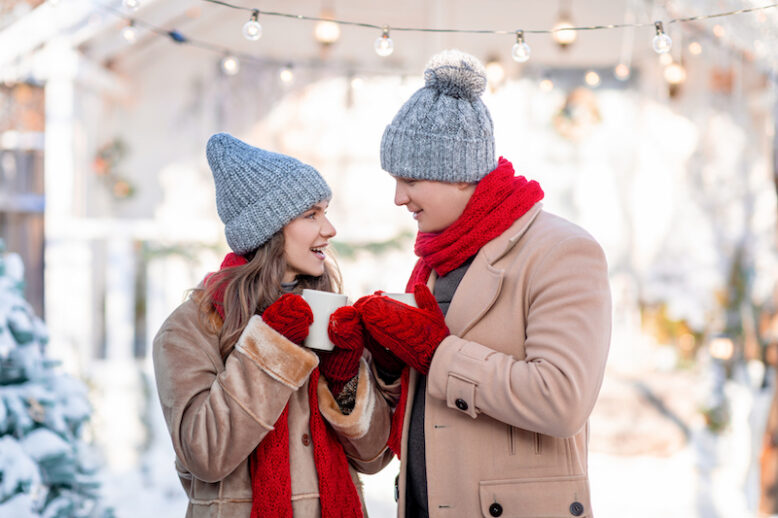 Beautiful lovers millennial man and woman posing together at snowy backyard, drinking hot tea, looking at each other and smiling, 