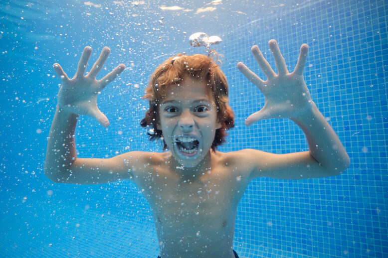 Boy with mouth opened screaming with eyes wide open in clear water of swimming pool and looking at camera