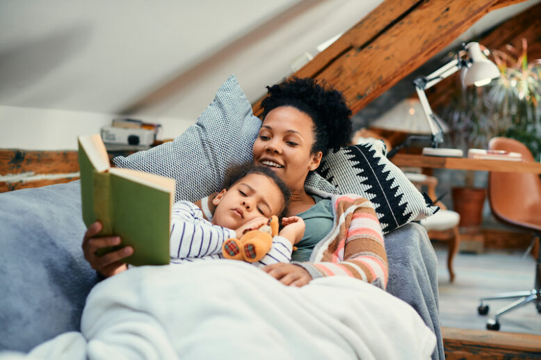 Little African American girl falls asleep while her mom reads a book on the couch at home.