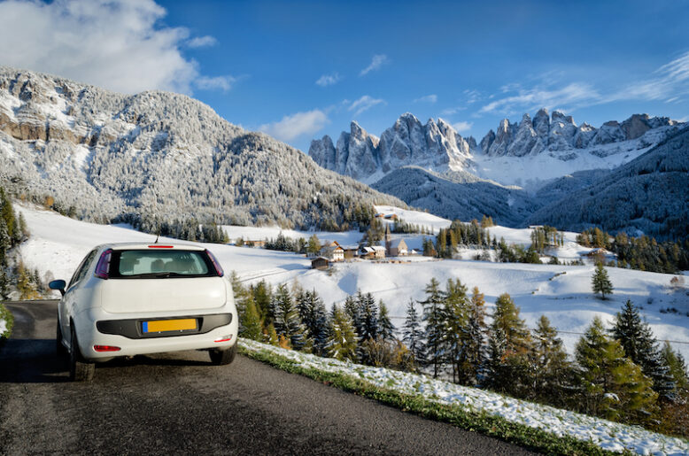 White car on the road during a road trip in a Dolomites landscape in winter with snow on the trees, the Geisler mountains and the church of the village St. Magdalena or Santa Maddalena in the Villnoesstal (Val di Funes valley) in South Tyrol, Italy.