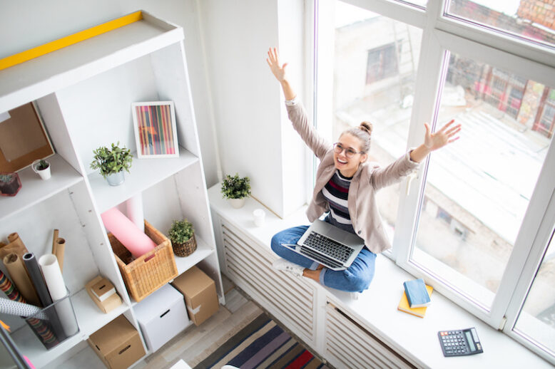 Ecstatic businesswoman with laptop raising her arms while sitting on windowsill and expressing victory