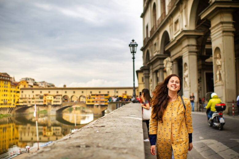 A tourist girl with an orange coat happily walks on the embankment in Florence, Italy.