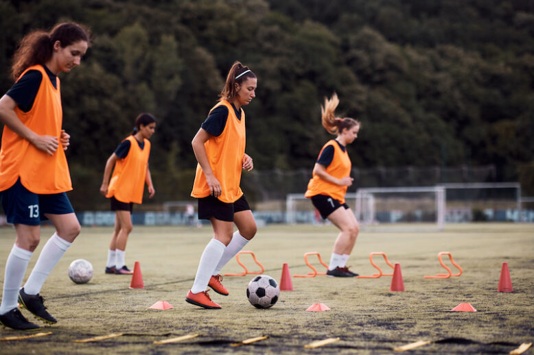 Female soccer player leading the ball among cones during sport training at the stadium.