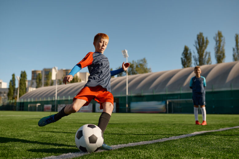 Boy wearing uniform playing football on field and kicking soccer ball for penalty