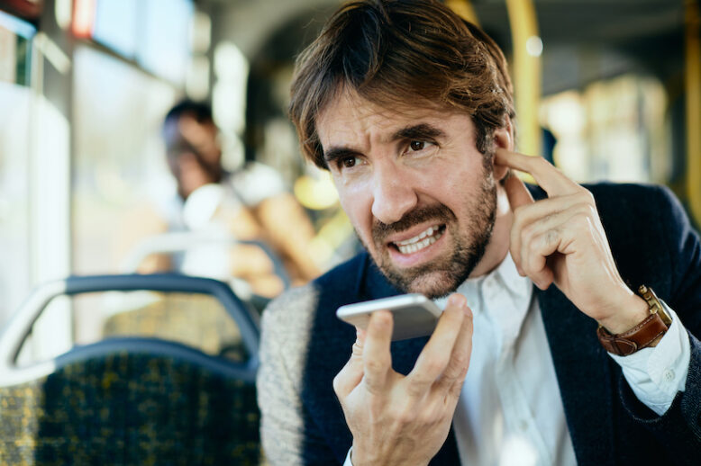 Businessman talking over mobile phone's speaker and holding finger in his ear while commuting by public transport during rush hour.