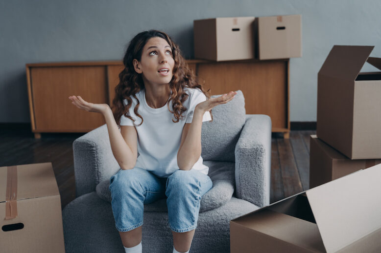 Displeased divorced young woman exclaiming shrugging sitting on armchair with cardboard boxes, tired after packing things for relocation. 