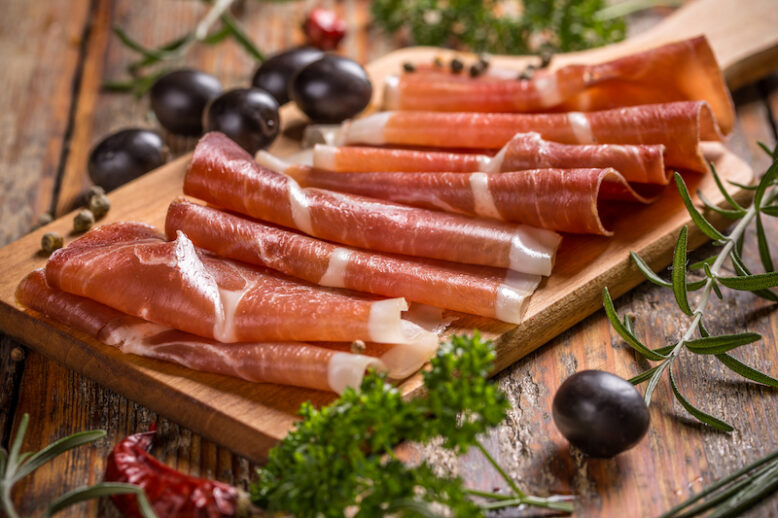 Thin slices of prosciutto with olives on wooden cutting board