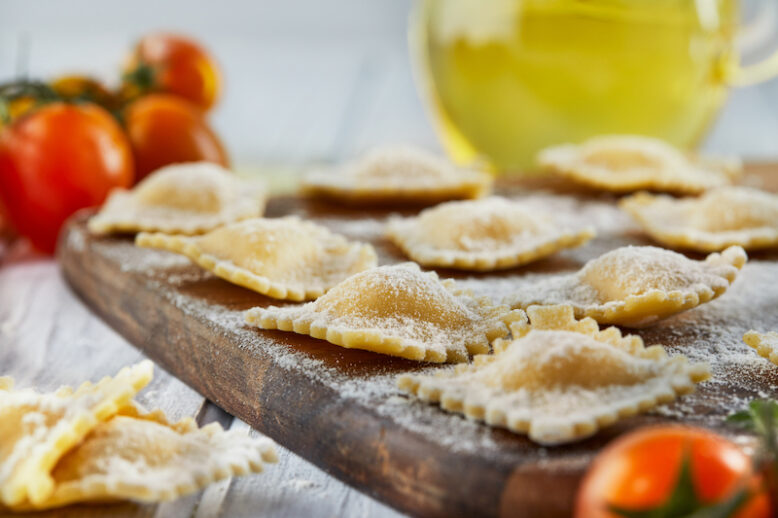 Tasty raw ravioli with flour, cherry tomatoes, sunflower oil and basil on a light wooden background. The process of making Italian ravioli.