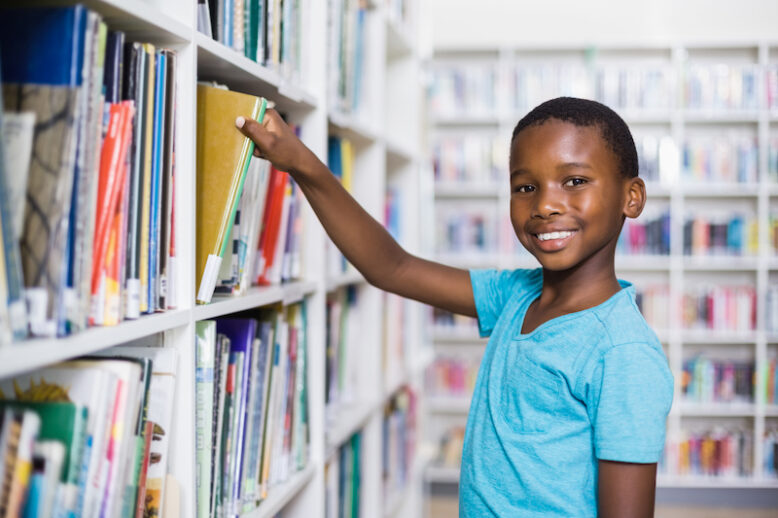 Portrait of schoolboy selecting a book from bookcase in library at school