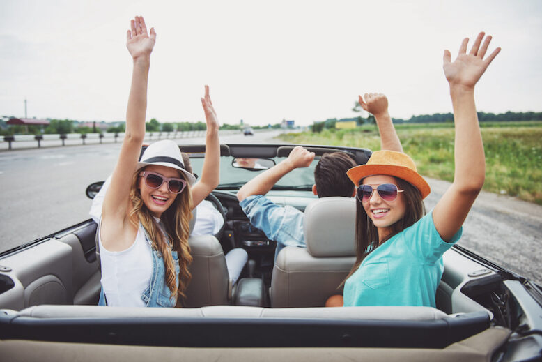 Shot back. Group of friends traveling on the cabriolet with hands up. Girls looking at camera.