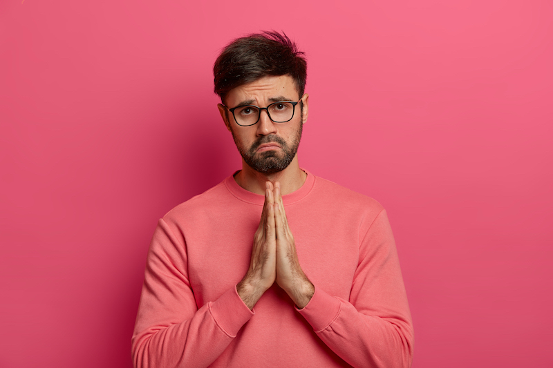 Desperate guilty unshaven man shows clasped hands, says sorry for bad mistake, beggs for help, looks unhappily at camera, wears spectacles and casual jumper, poses indoor over rosy background.