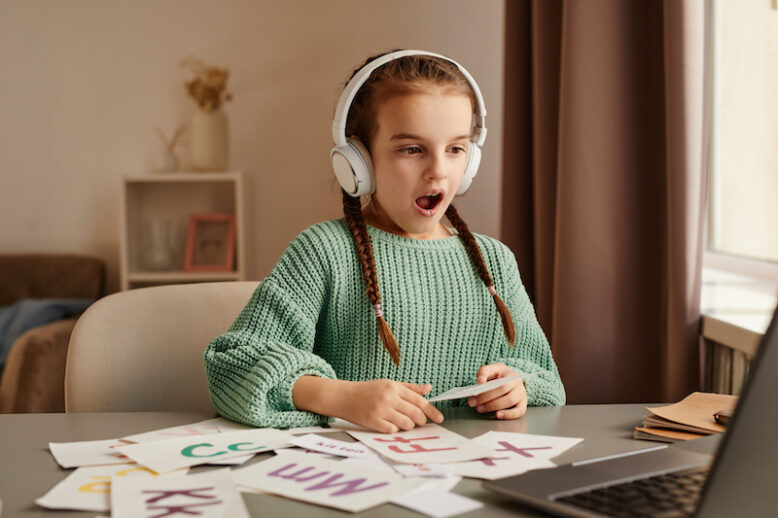 Little girl in wireless headphones sitting at the table and learning to pronounce new english words during online lesson on laptop