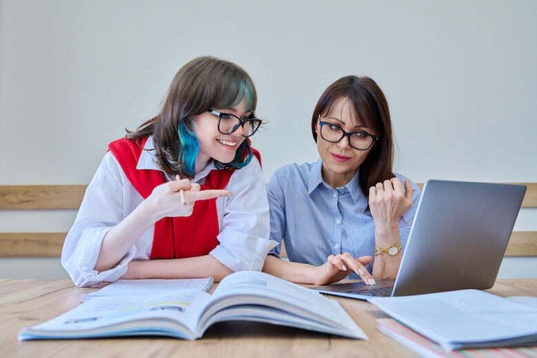 Young teenage female studying foreign languages in course with teacher. Tutor and student sitting at desk in office, using books, notebooks, laptop computer. Education, knowledge, learning concept