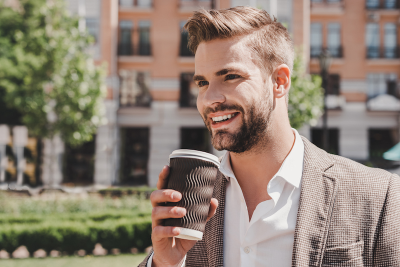 Young stylish man wearing brown jacket, white shirt and sunglasses outdoors standing on the city street with a cup of coffee looking aside and smiling joyful.