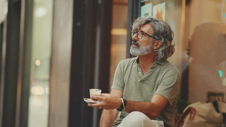 Close-up, thoughtful middle-aged man with gray hair and beard, wearing casual clothes, sits in street cafe. Mature gentleman in eyeglasses drinks aroma coffee