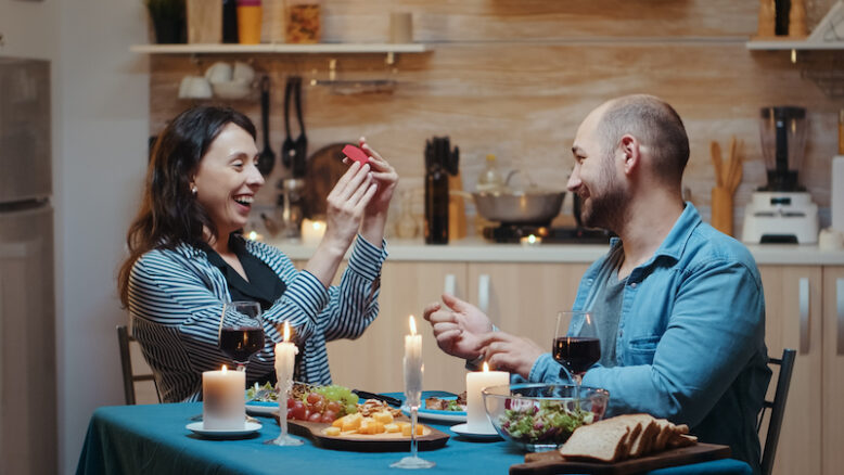Handsome man proposing to his girlfriend marriage during festive dinner, in kitchen sitting at the table drinking a glass of red wine.