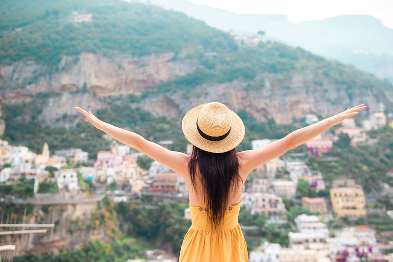 Rear view of young woman wearing straw hat and yellow dress with Positano village in the background, Amalfi Coast, Italy
