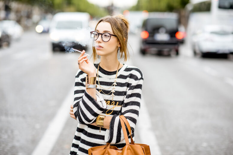 Young stylish woman in striped sweater with eyeglasses smoking a cigarette standing outdoors on the street in Paris