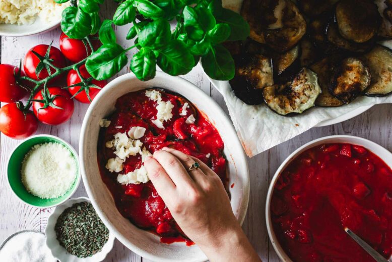 Sprinkling mozzarella on top of tomato sauce in a casserole dish, surrounded by tomatoes, parmesan, oregano, canned tomatoes, fried eggplant, basil plant