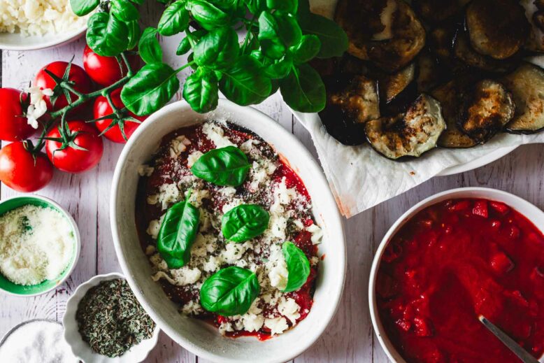 Fresh basil on top of tomato sauce in a casserole dish, surrounded by tomatoes, parmesan, oregano, canned tomatoes, fried eggplant, basil plant
