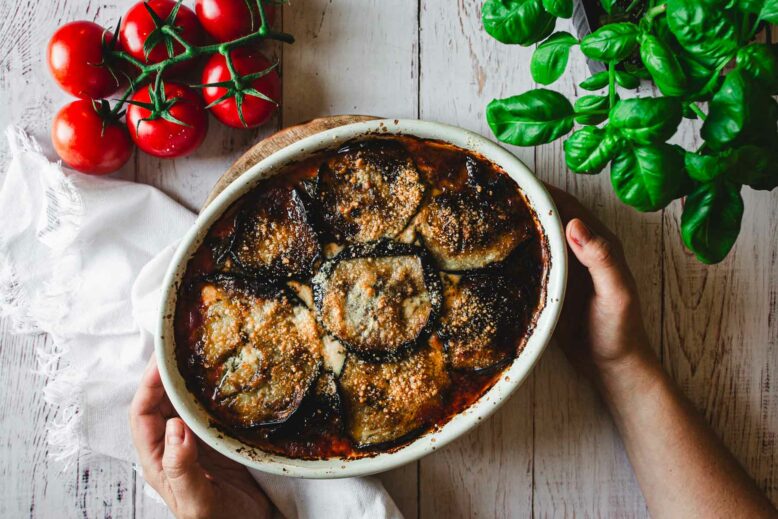 Hands serving baked Eggplant Parmiagana with white napkin, red tomatoes and green basil