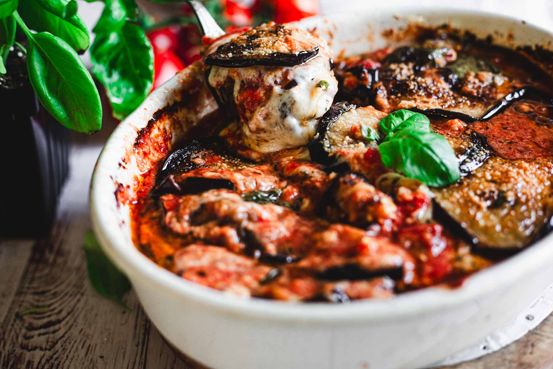 Lifting a spoon of baked Eggplant Parmiagana with basil on top of white napkin, red tomatoes and green basil