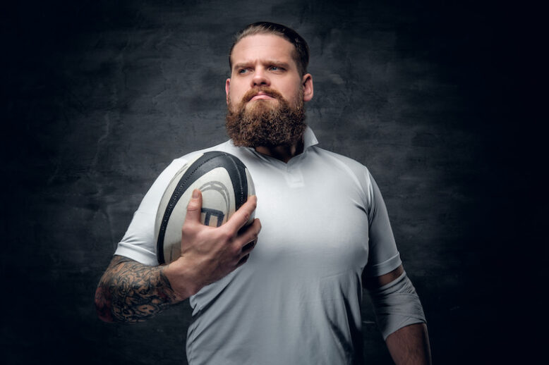 Portrait of bearded rugby player with tattoos on his arms holds a game ball.