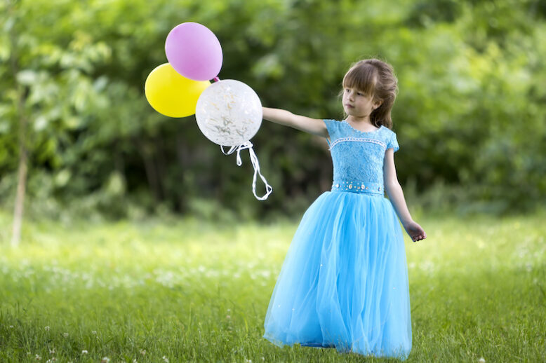 Beautiful little blond long-haired girl in nice long blue evening dress looking like princess looks in camera holding colorful balloons standing in blooming field on blurred green trees background.