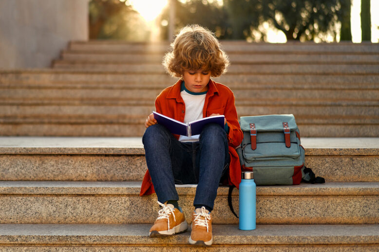 Child with a backpack sits on the stairs near the school and reads a textbook.