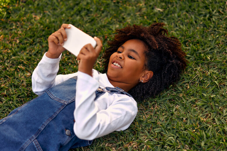 Adorable stylish african american school age girl lying on the grass in the park, relaxing and playing games on the phone, having fun on the day off. School break.