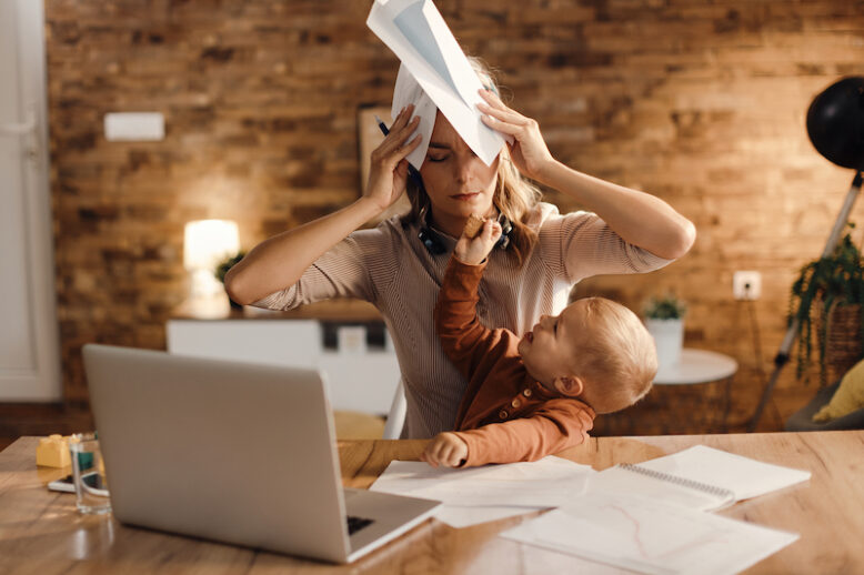 Stressed working mother covers her head with paperwork while her young son gives her a cookie at home.