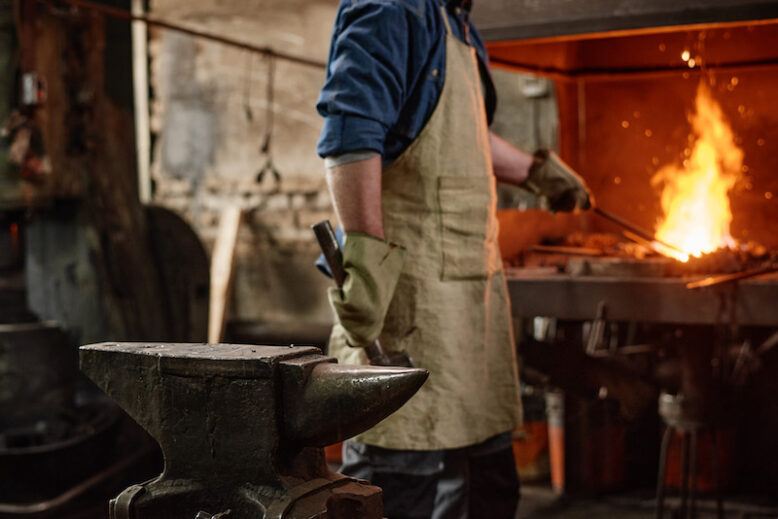 Close-up of worker adding coal in furnace while working with hammer in the blacksmith shop