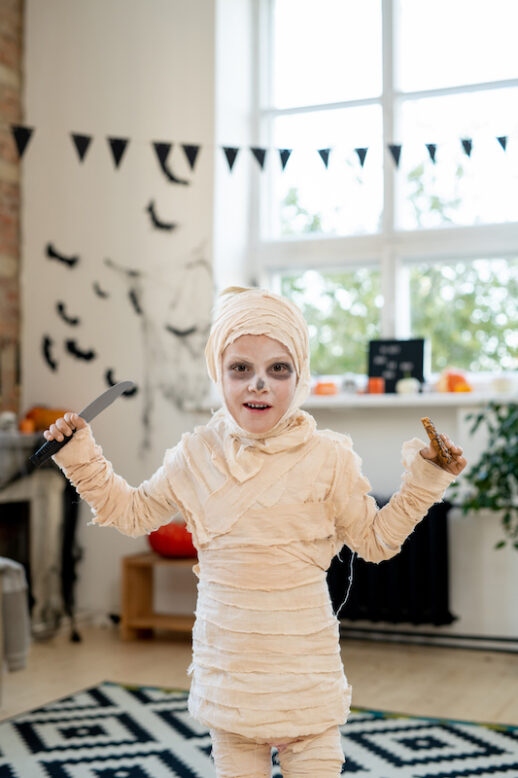 Portrait of cute boy in mummy costume standing with knife and cookie as symbol of trick-or-treating tradition for Halloween