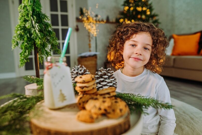 Happy little boy with curly hair waiting for Santa Claus prepairing milk and cookies at Christmas time at home.