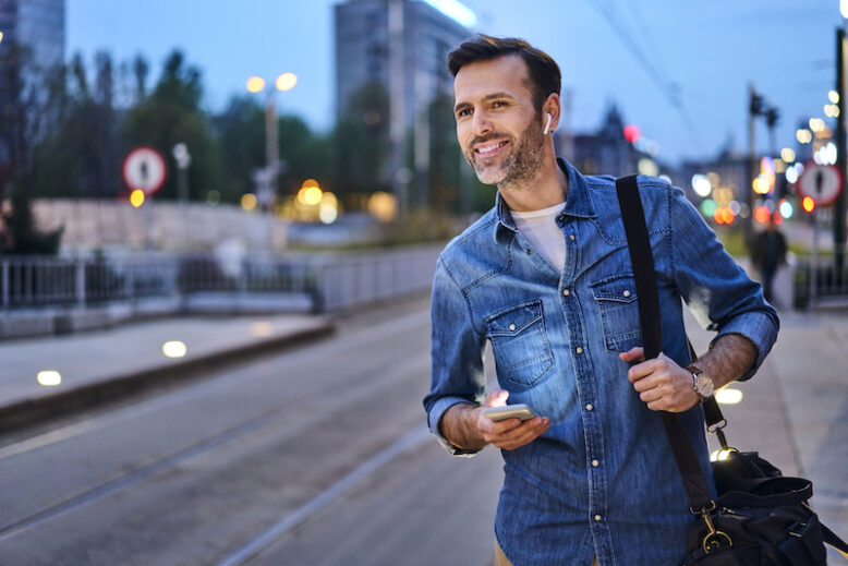 Smiling man wearing wireless headphones and smartphone waits at tram stop during evening commute.