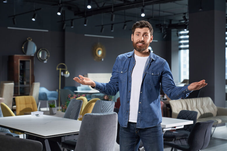 Strong bearded retailer shrugging, confused expression with arms and hands raised, while working. Portrait of young man in casual clothes making doubt gesture in furniture store. Concept of doubt.