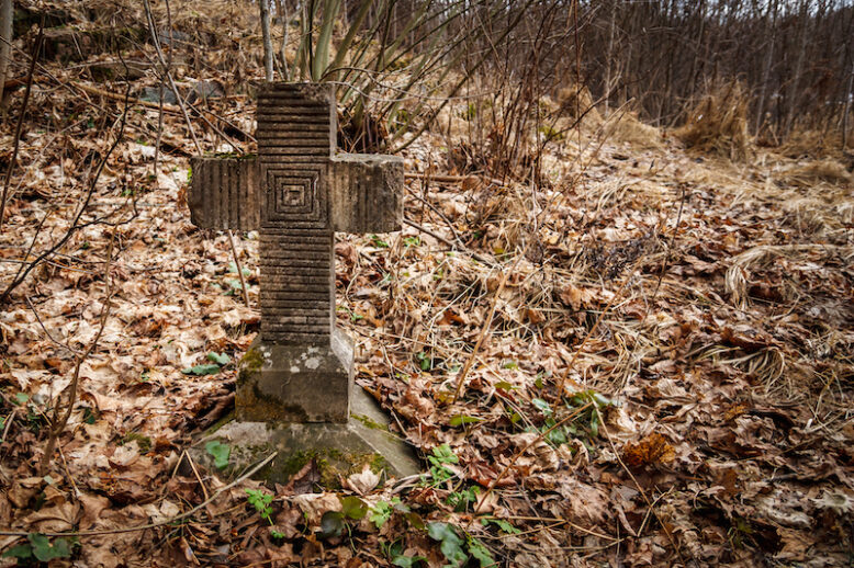 Old tombstone cross overgrown with moss and old leaves in the autumn forest