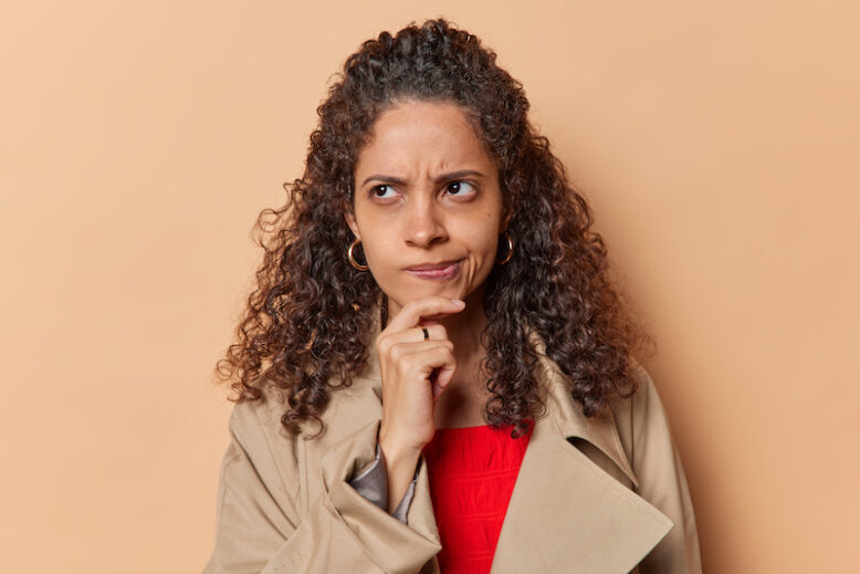 Unsure displeased Afro American purses lips keeps hand on chin frowns face and ponders on something looks doubtfully dressed in jacket isolated over brown background feels unaware or questioned